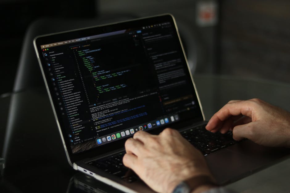 A close-up view of a person's hands typing on a laptop keyboard, with the screen displaying lines of code in a dark-themed text editor. The laptop is positioned on a black surface, and the background 
